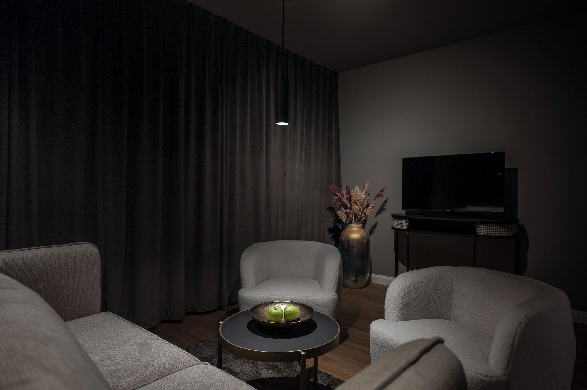 A modern living room with a sofa, armchairs, a coffee table with apples, a vase with dried plants, and a TV on a stand.