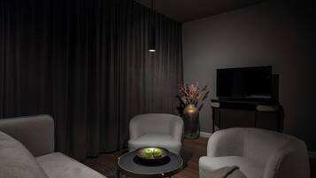 A modern living room with a sofa, armchairs, a coffee table with apples, a vase with dried plants, and a TV on a stand.