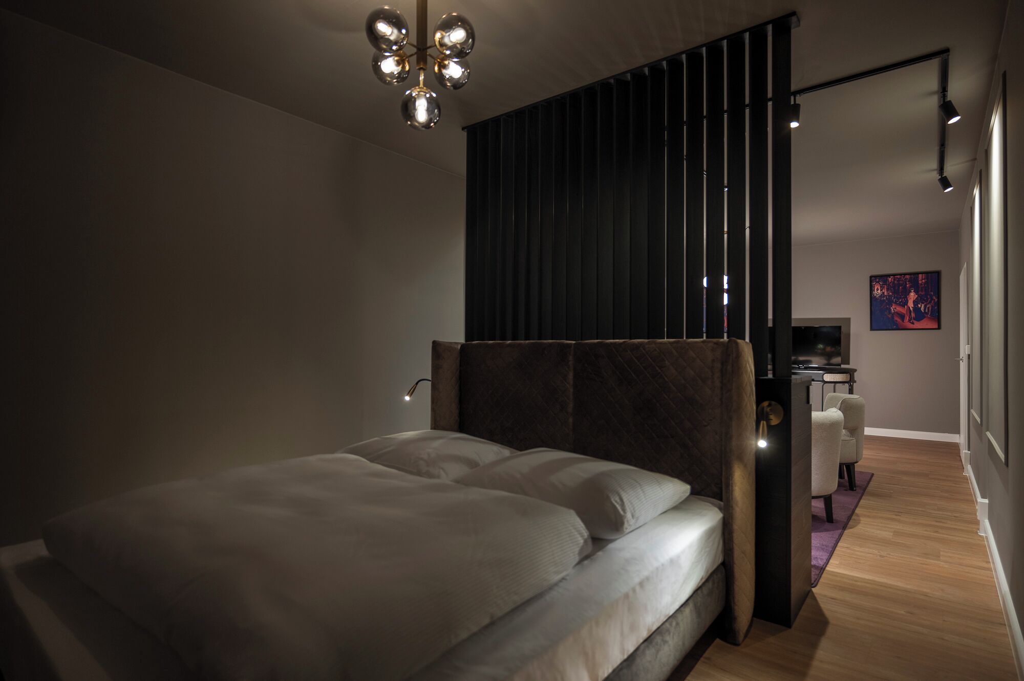 A modern bedroom with a large bed, a dark partition, a desk with a computer, and a TV.
