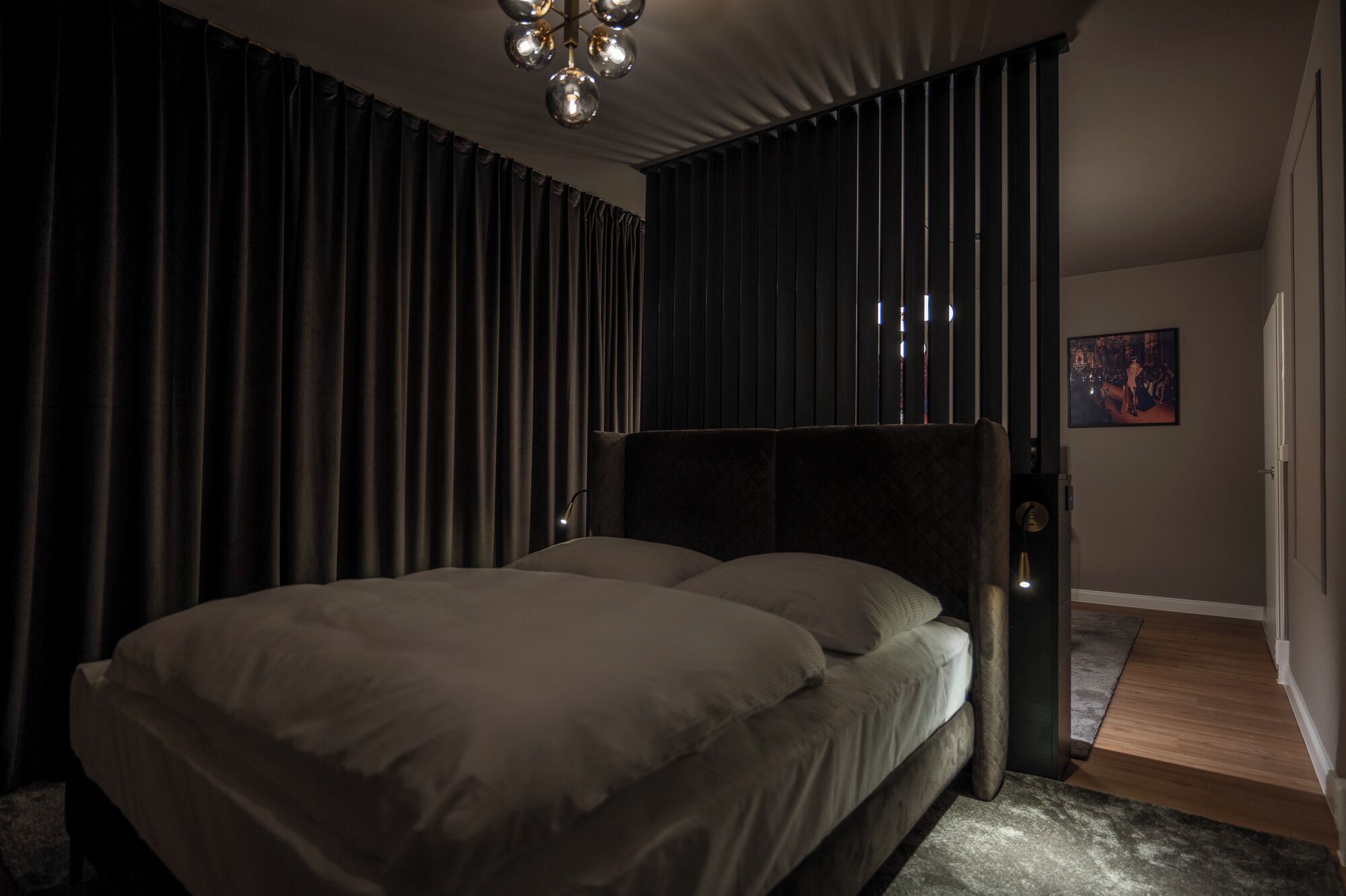 Een moderne slaapkamer met een groot bed, donkere gordijnen en een kroonluchter.
