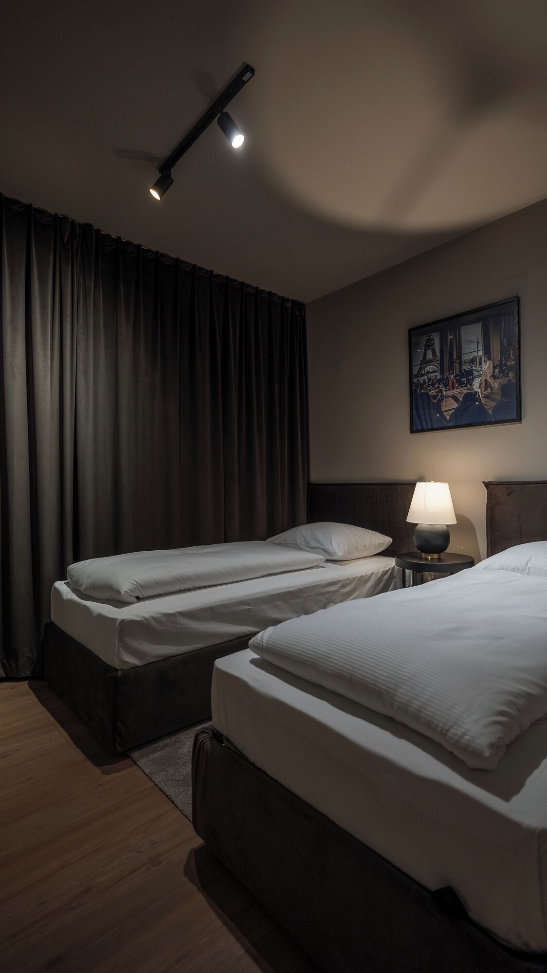 A hotel room with two single beds, a nightstand with a lamp, a painting of the Eiffel Tower, and a large window with curtains.