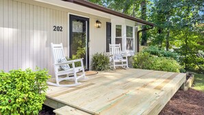 A wooden deck with two rocking chairs and a bench, a door with the number 202, and a small garden area with plants and a bench.