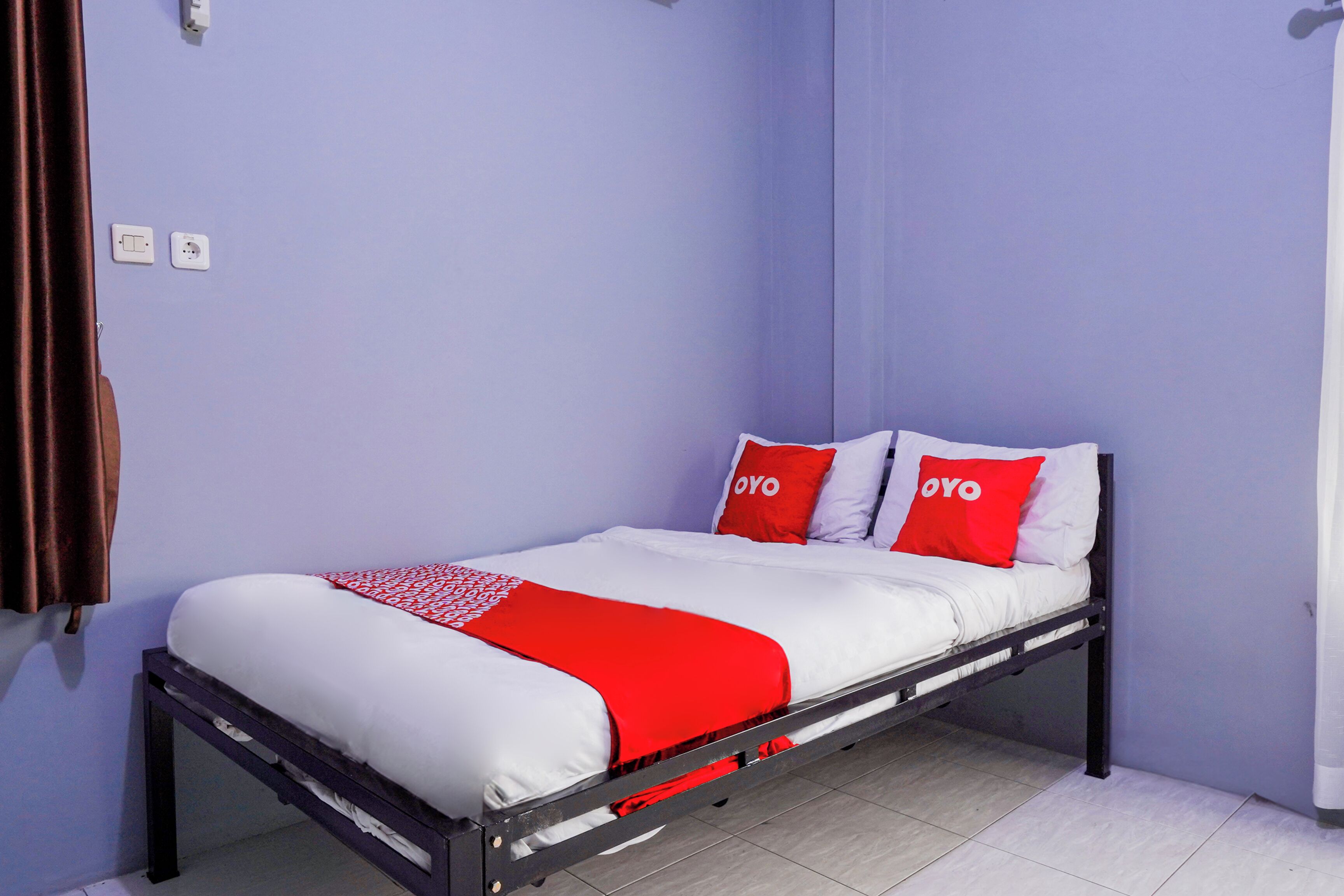 Uma cama de solteiro com colcha vermelha e branca, duas almofadas vermelhas com a palavra "OYO" estampada e uma cortina marrom no lado esquerdo.