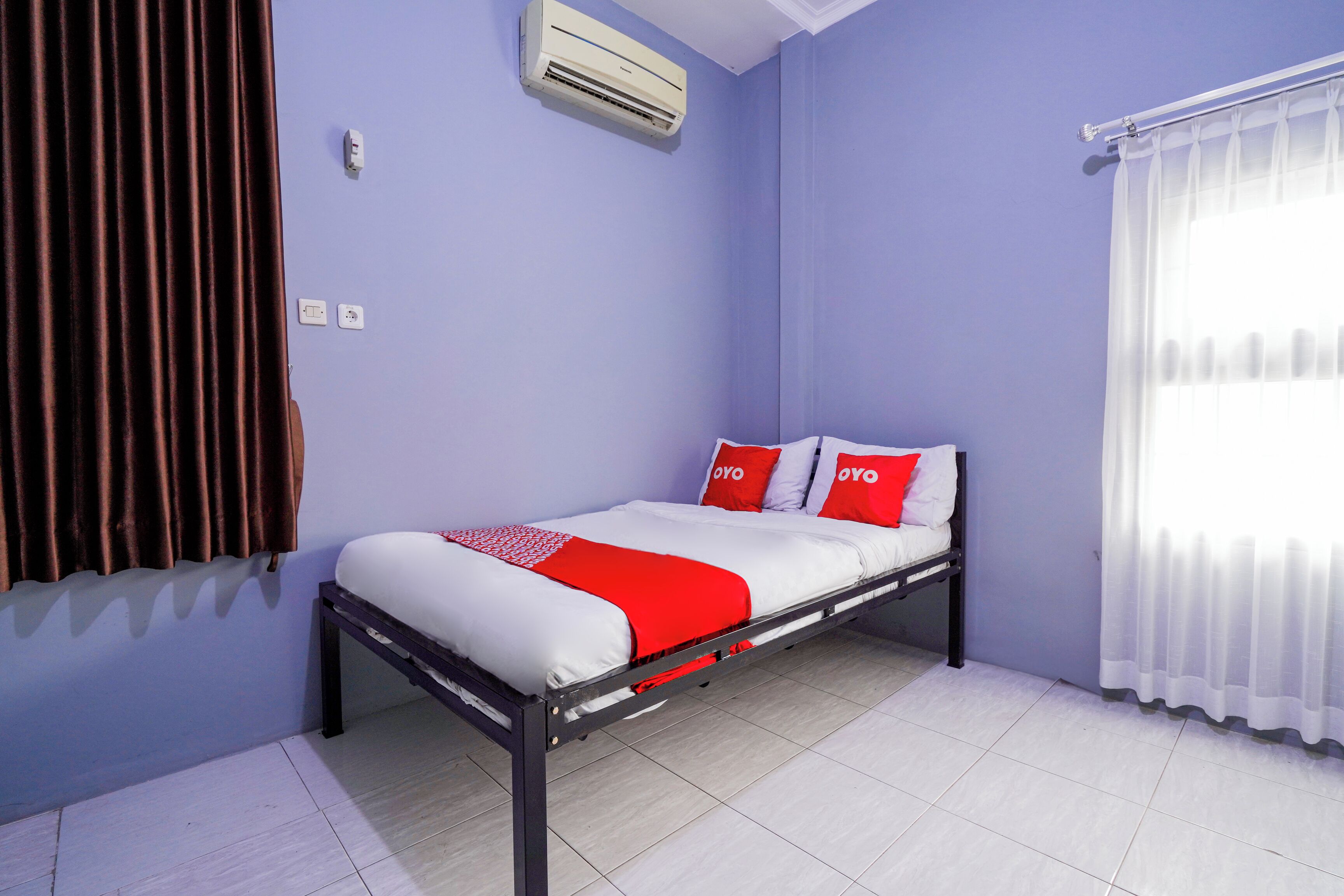 A single bed with a white and red bedspread, two pillows with the brand name "OYO," a wall-mounted air conditioner, and a window with white curtains.