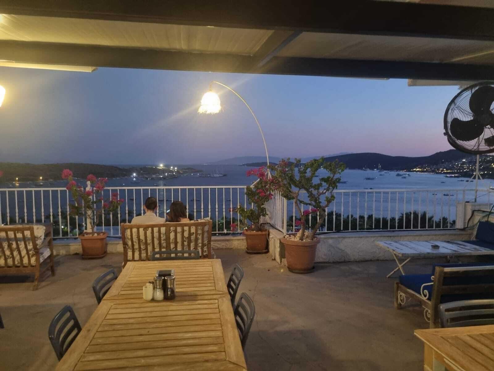 Un área de estar al aire libre con vistas al mar y barcos. Se observan mesas y sillas de madera, plantas en macetas y un ventilador.