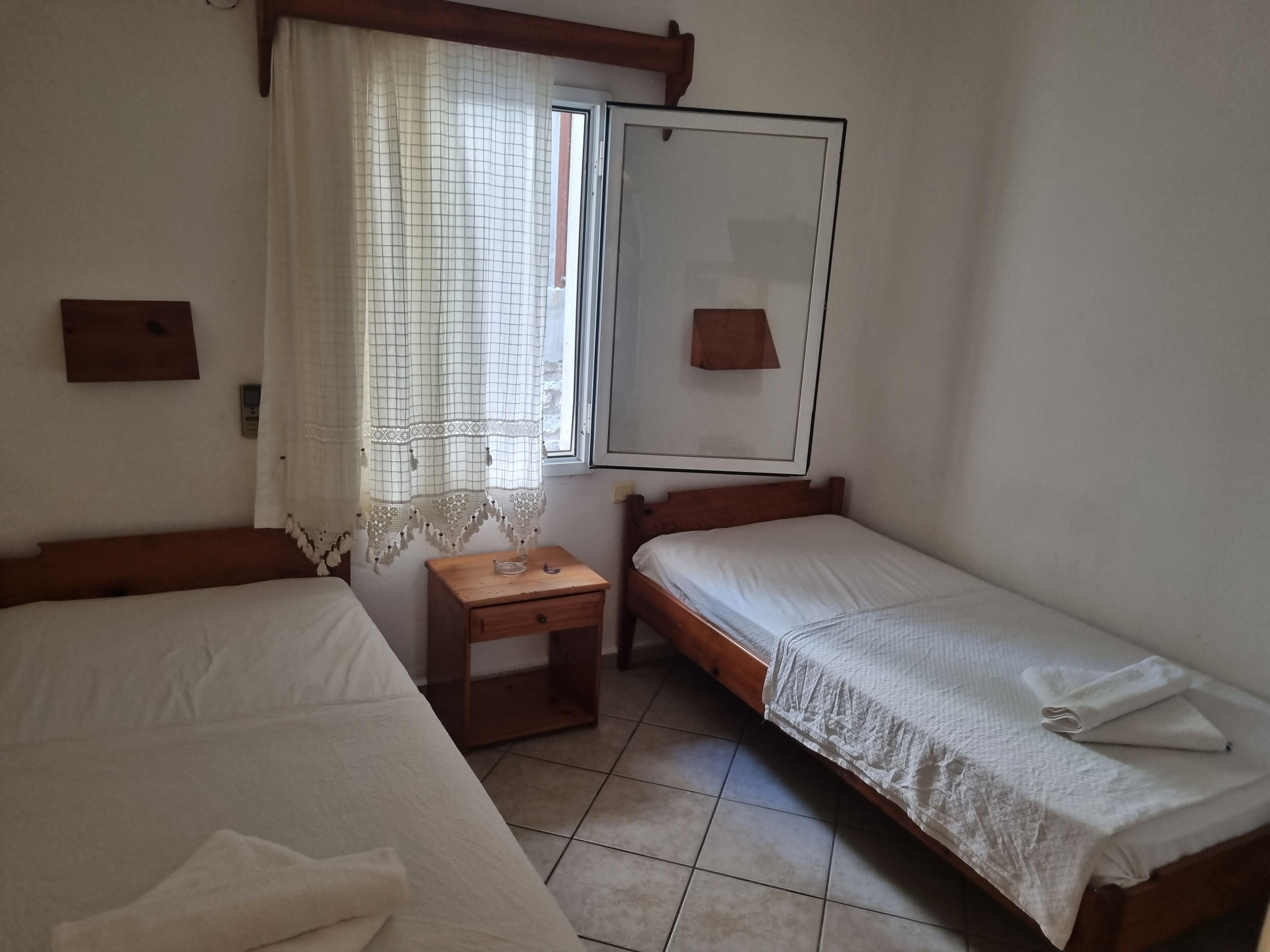 Een kamer met twee enkele bedden, een klein houten nachtkastje, een raam met gordijn en een spiegel aan de muur.