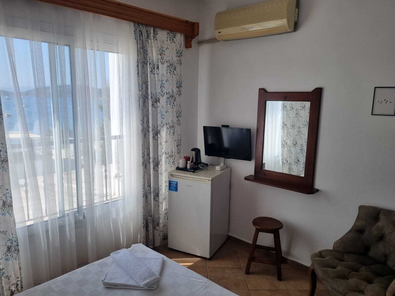 A hotel room with a bed, a desk with a television, a mirror, and a window with sheer curtains.
