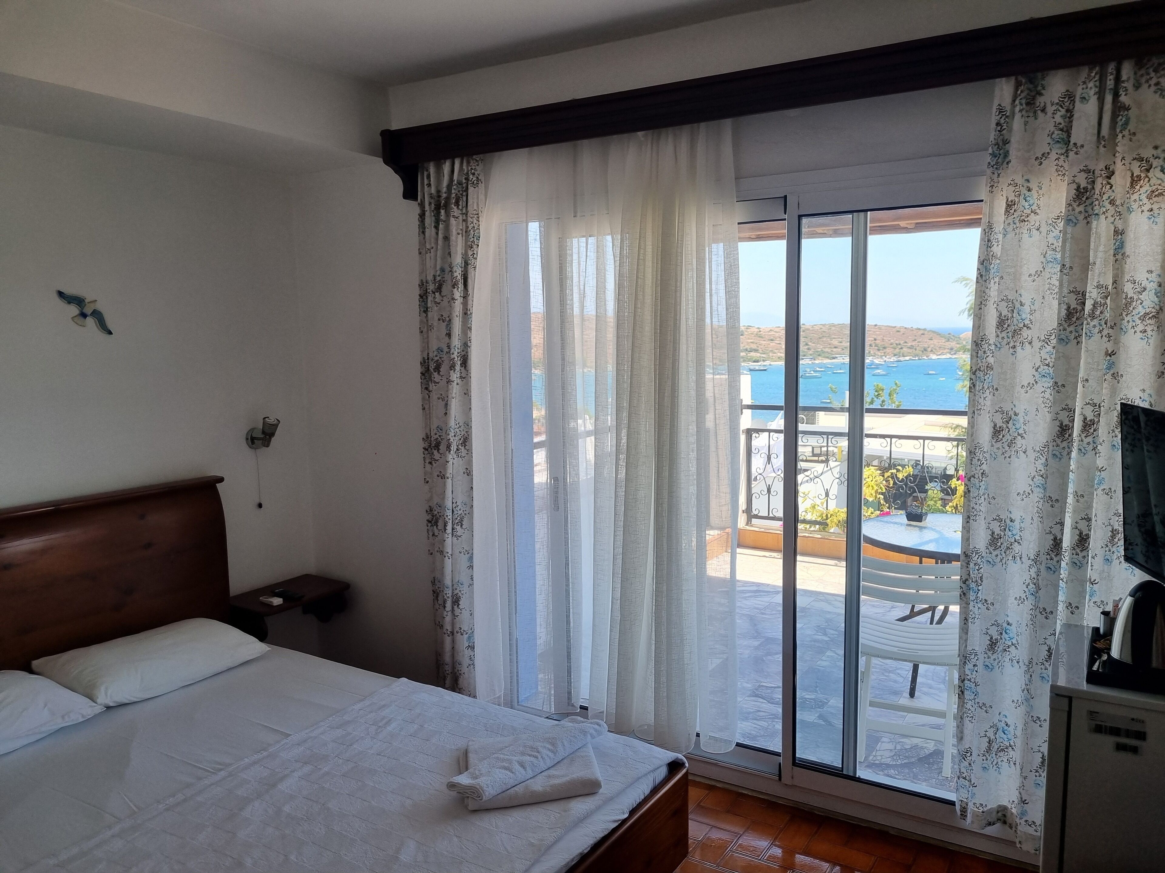 A hotel room with a bed, a nightstand, a TV, and a balcony with a view of the sea.