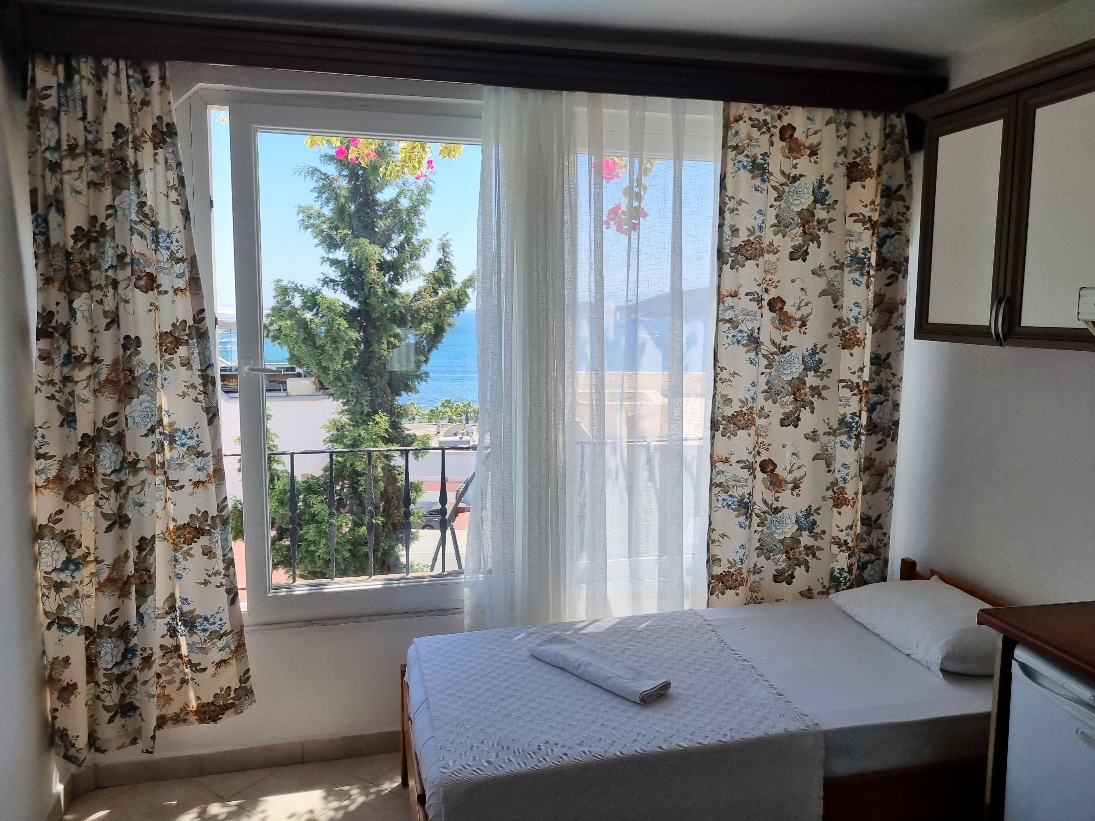A hotel room with a bed, a window with floral curtains, and a view of the sea and trees.