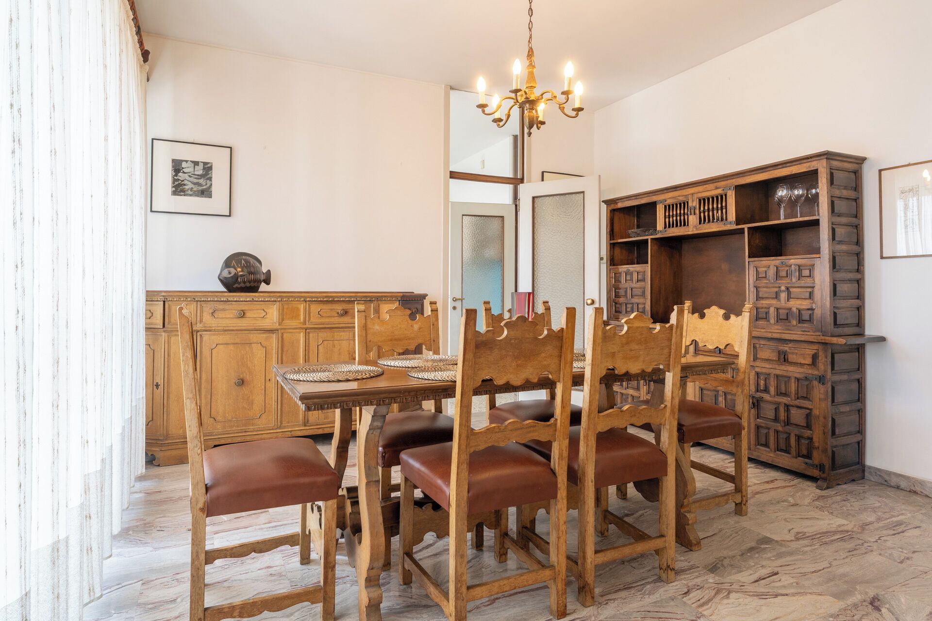 Uma sala de jantar com uma mesa e cadeiras de madeira, um lustre e um armário de madeira com prateleiras.