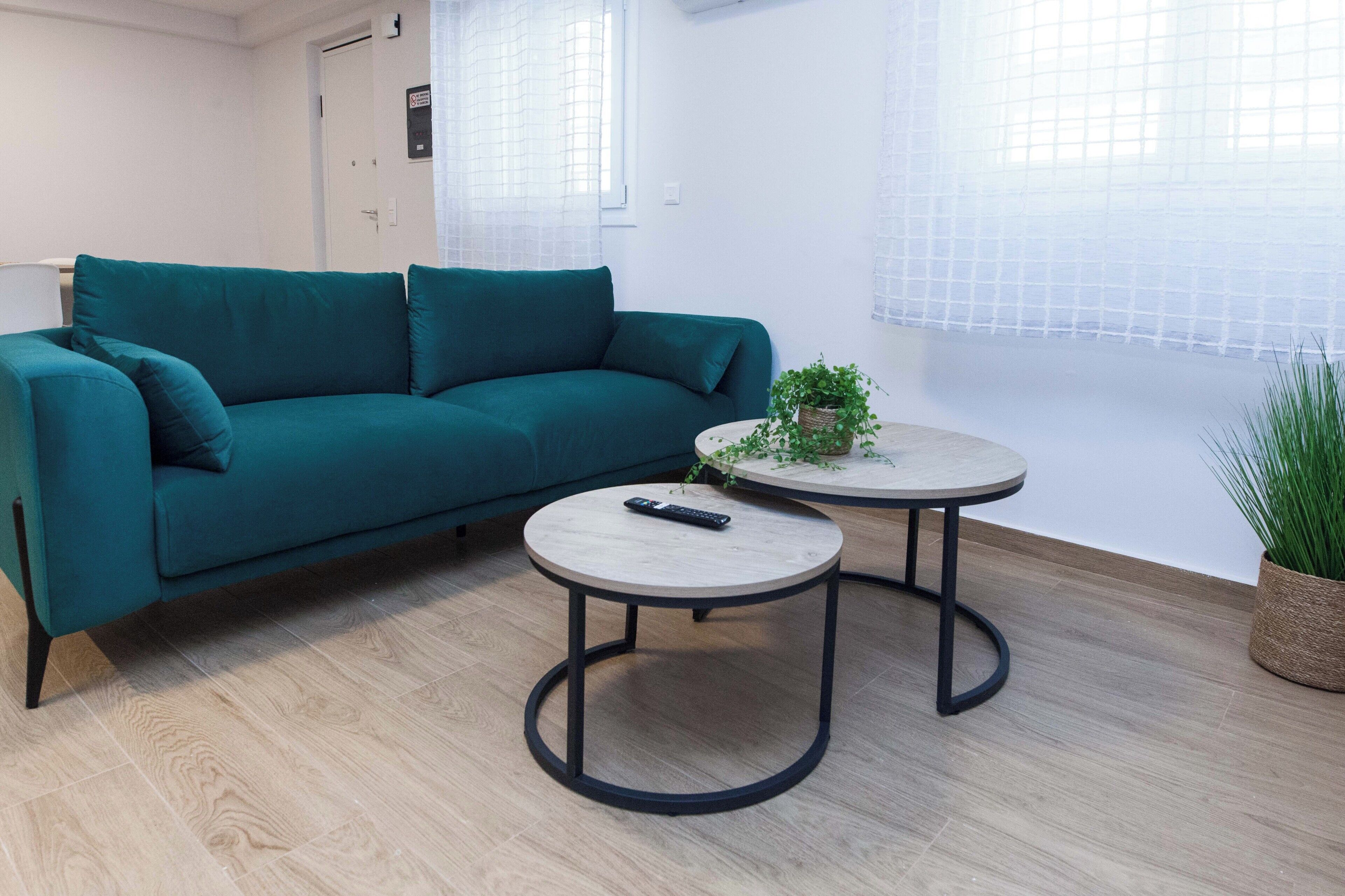 Un sofá color turquesa, dos mesas redondas y una planta en maceta en una habitación con piso de madera clara y paredes blancas.