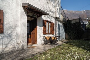Un bâtiment rustique avec une porte et des fenêtres en bois, un petit espace extérieur aménagé avec une table et des chaises, et une vue sur les montagnes en arrière-plan.