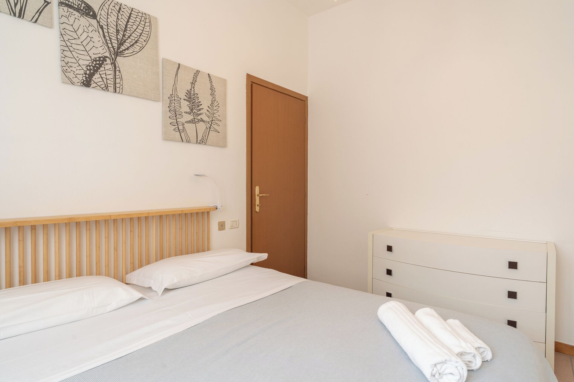 Un dormitorio con cama, cabecera de madera, pared blanca con obra de arte, una puerta y una mesita de noche blanca con toallas.