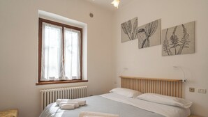 A bedroom with a bed, a window with curtains, a radiator, and three wall-mounted plant illustrations.