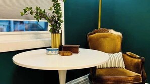 Una habitación con una mesa redonda blanca, una silla tapizada en verde, una lámpara de pie y un ventanal con persianas.