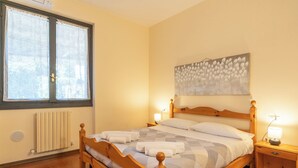 A bedroom with a wooden bed, a radiator, a window with curtains, a painting on the wall, and a bedside table with a lamp. - SunsetView - Lake Como swimming pool and garden (Lierna)