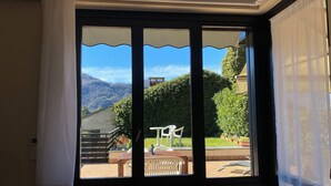A room with a large window overlooking a patio area with outdoor furniture and a view of mountains. - SunsetView - Lake Como swimming pool and garden (Lierna)