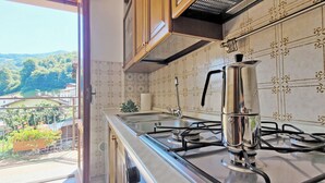 Une cuisine équipée d’une cuisinière à gaz, d’une cafetière et offrant une vue sur un paysage verdoyant par la fenêtre.