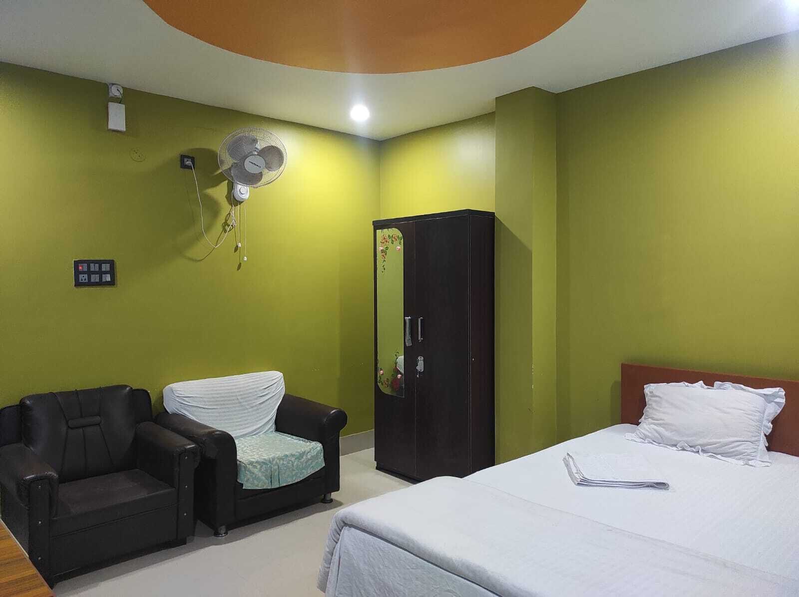 A hotel room with a bed, a black leather chair, a wardrobe, and a wall-mounted fan.