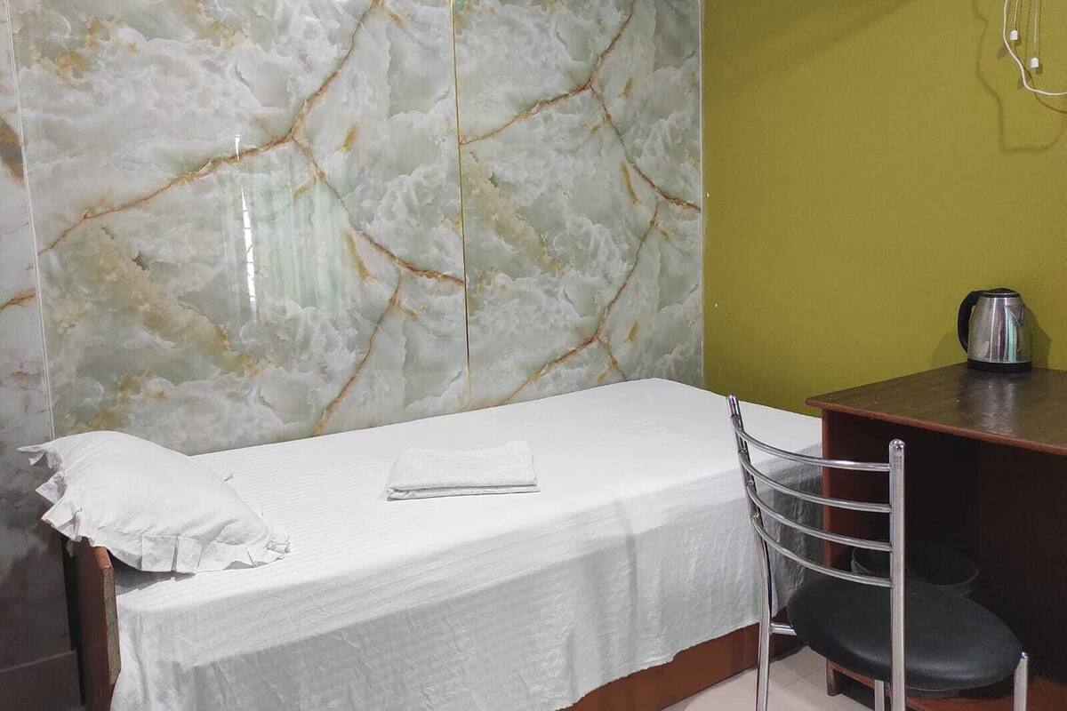 A single bed with a white sheet, a metal chair, a small table with a kettle, and a marble-patterned wall.