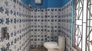 A small bathroom with a toilet, a shower, and a window with bars. - Raj Hotel (Baksa)