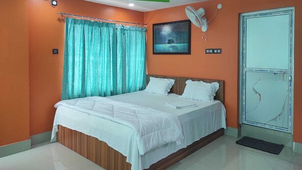 A bedroom with a bed, curtains, a fan, and a wall-mounted picture. - Raj Hotel (Baksa)