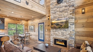 A log cabin interior with a stone fireplace, wooden furniture, and a dining area with a view of the outdoors. - 2 Bedroom w/ Fire Pit, Indoor/Outdoor Fireplace, Hot Tub, Cornhole, Shuffleboard (Sevierville)