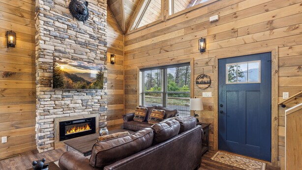 A log cabin interior with a stone fireplace, leather sofa, and a blue door. - 2 Bedroom w/ Fire Pit, Indoor/Outdoor Fireplace, Hot Tub, Cornhole, Shuffleboard (Sevierville)