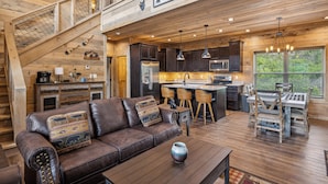 A spacious cabin interior with a high ceiling, wooden walls, a kitchen area, a dining space, and a cozy seating area with a leather couch and a coffee table. - 2 Bedroom w/ Fire Pit, Indoor/Outdoor Fireplace, Hot Tub, Cornhole, Shuffleboard (Sevierville)