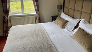 Ein ordentlich bezogenes Bett mit einem gemusterten Überwurf, einem Kopfteil und zwei Kissen. Ein Fenster mit Vorhängen ist sichtbar, und ein Nachttisch mit einer Lampe steht daneben.
