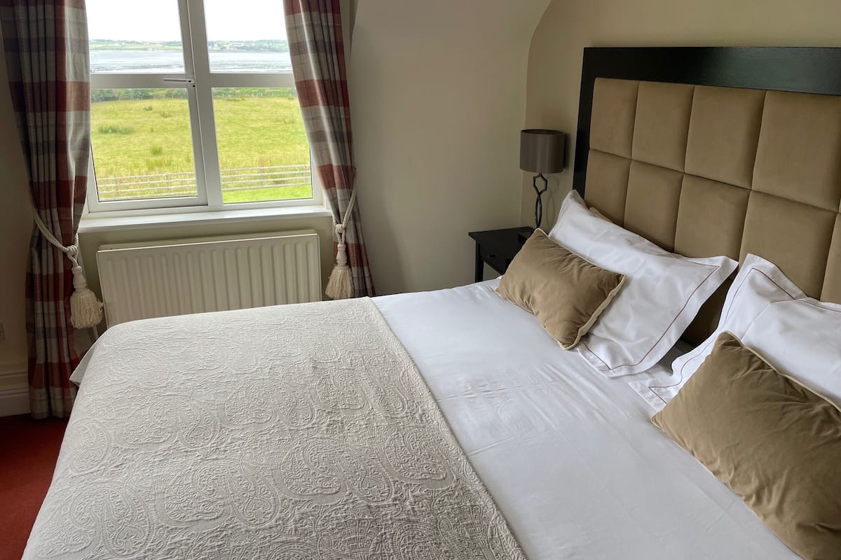 A neatly made bed with a patterned bedspread, a headboard, and two pillows. A window with curtains is visible, and a nightstand with a lamp is next to the bed.