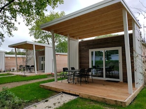 A modern outdoor patio area with a wooden deck, metal-framed tables and chairs, and a glass door leading to a covered space. - Nice Chalet With Airco Near Efteling (Kaatsheuvel)