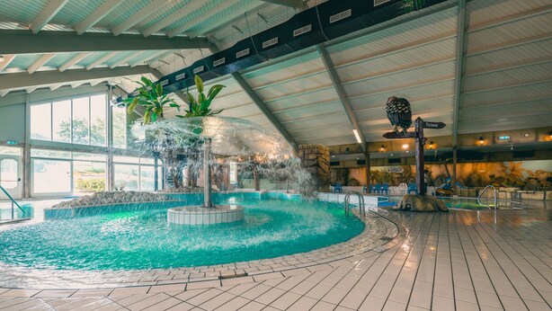 Een binnenzwembad met een centraal waterkunstwerk, omringd door een betegelde vloer en een hoog plafond met dakramen.