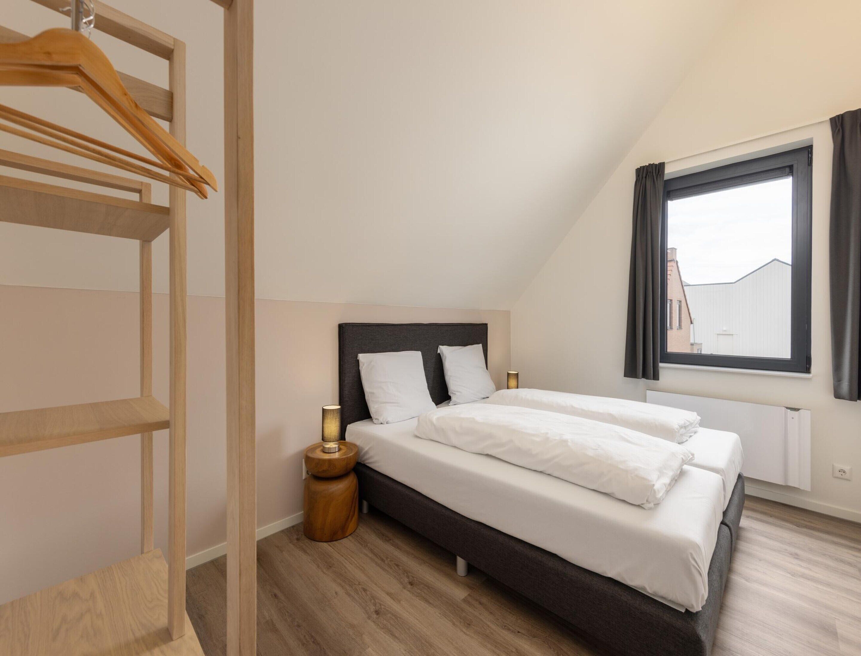 Ein Schlafzimmer mit einem Bett, einem Holzschrank, einem Nachttisch, einer Lampe und einem Fenster mit Vorhängen.