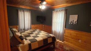 A bedroom with a wooden ceiling, a bed with a checkered blanket, a television on the wall, and a red floral rug. - Welcome Outdoor Enthusiasts! Direct ATV access.  (Bingham)