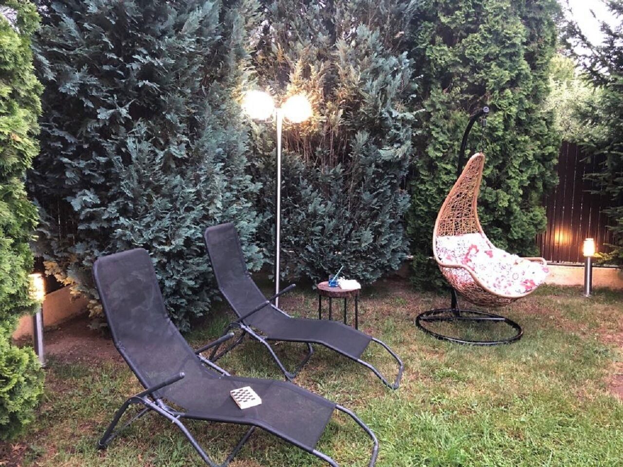 Two lounge chairs, a small table, and a decorative chair in a garden area with dense foliage and a street lamp.
