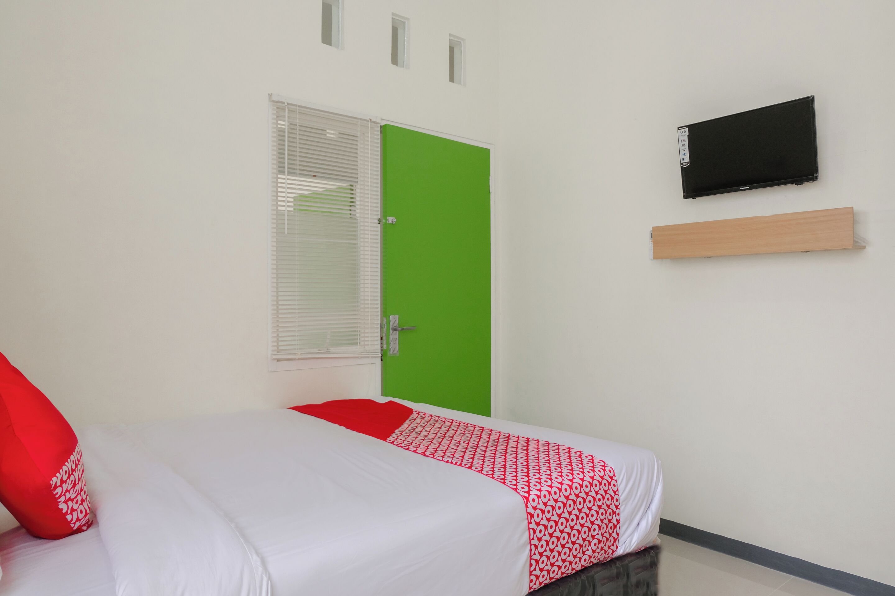 A room with a bed, a green door, a TV mounted on the wall, and a window with blinds.
