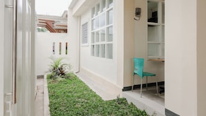 Un estrecho pasillo exterior con un pequeño jardín, una silla color turquesa y una puerta blanca.