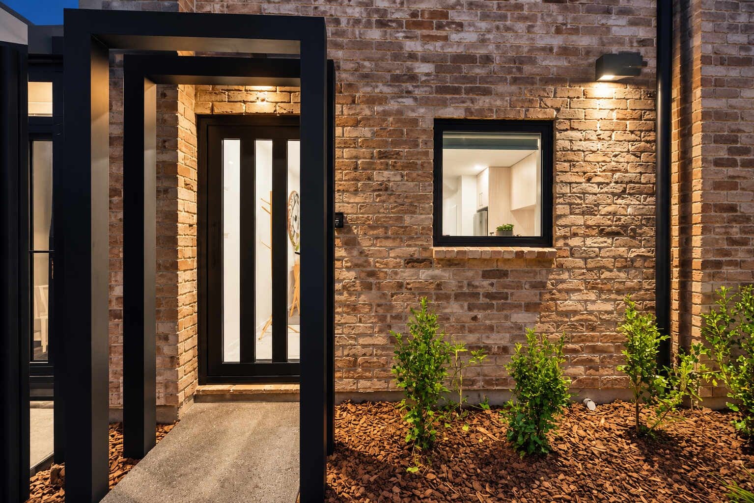Una casa moderna con fachada de ladrillo, una puerta de marco negro y un ventanal con luminaria.