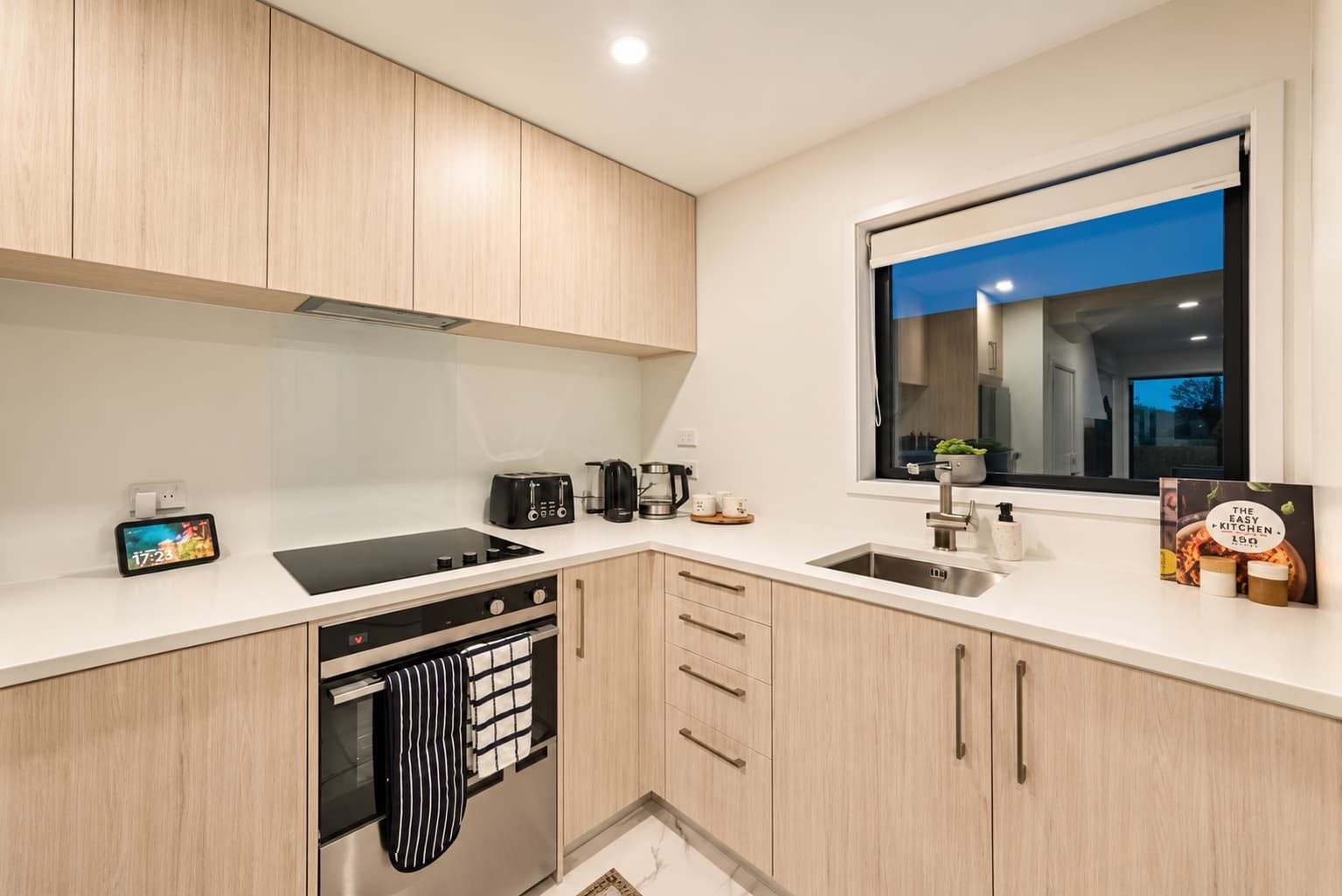Una cocina moderna con armarios de madera clara, una cocina de inducción negra, una cafetera, un tostador y un ventanal con vistas al exterior.