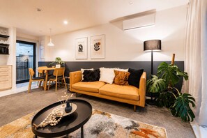Un salon moderne avec un canapé, une table basse et un coin repas avec des chaises. Deux œuvres d’art encadrées ornent le mur, ainsi qu’une plante en pot et une lampe sur pied.