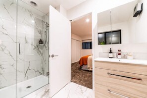 A modern bathroom with a glass shower enclosure, a white vanity with a mirror, and a bed in the background.
