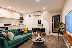 A modern living room with a green sofa, a dining table with chairs, a coffee table, and a TV mounted on the wall. - Central Stay – Sleeps 5 | Walk to Te Pae & Casino (Christchurch)