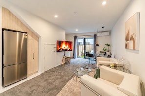 A modern living room with a large flat-screen TV, a built-in stainless steel refrigerator, and a cozy seating area with a sofa and a coffee table. - CBD 2BR Retreat | Private Garden + BBQ (Christchurch)
