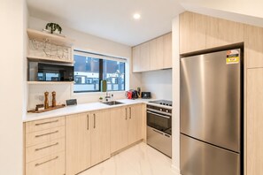 A modern kitchen with wooden cabinets, a stainless steel refrigerator, a built-in oven, and a window with a view of buildings. - CBD 2BR Retreat | Private Garden + BBQ (Christchurch)