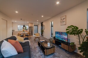 Una sala de estar moderna con sofá, mesa de centro y televisor. Se observa un comedor con sillas y mesa al fondo.