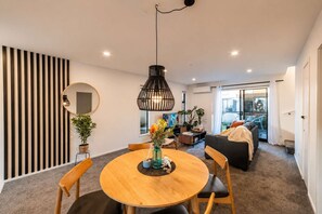 A modern living room with a round wooden table, a black hanging light fixture, a sofa, and a balcony with a view. - Merivale 2BR | Parking + Fenced Backyard (Christchurch)