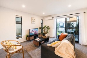 A modern living room with a flat-screen TV, a sofa, a coffee table, and a large window offering a view of the outdoors.