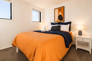 Un dormitorio con cama, mesita de noche, lámpara y un cuadro en la pared.