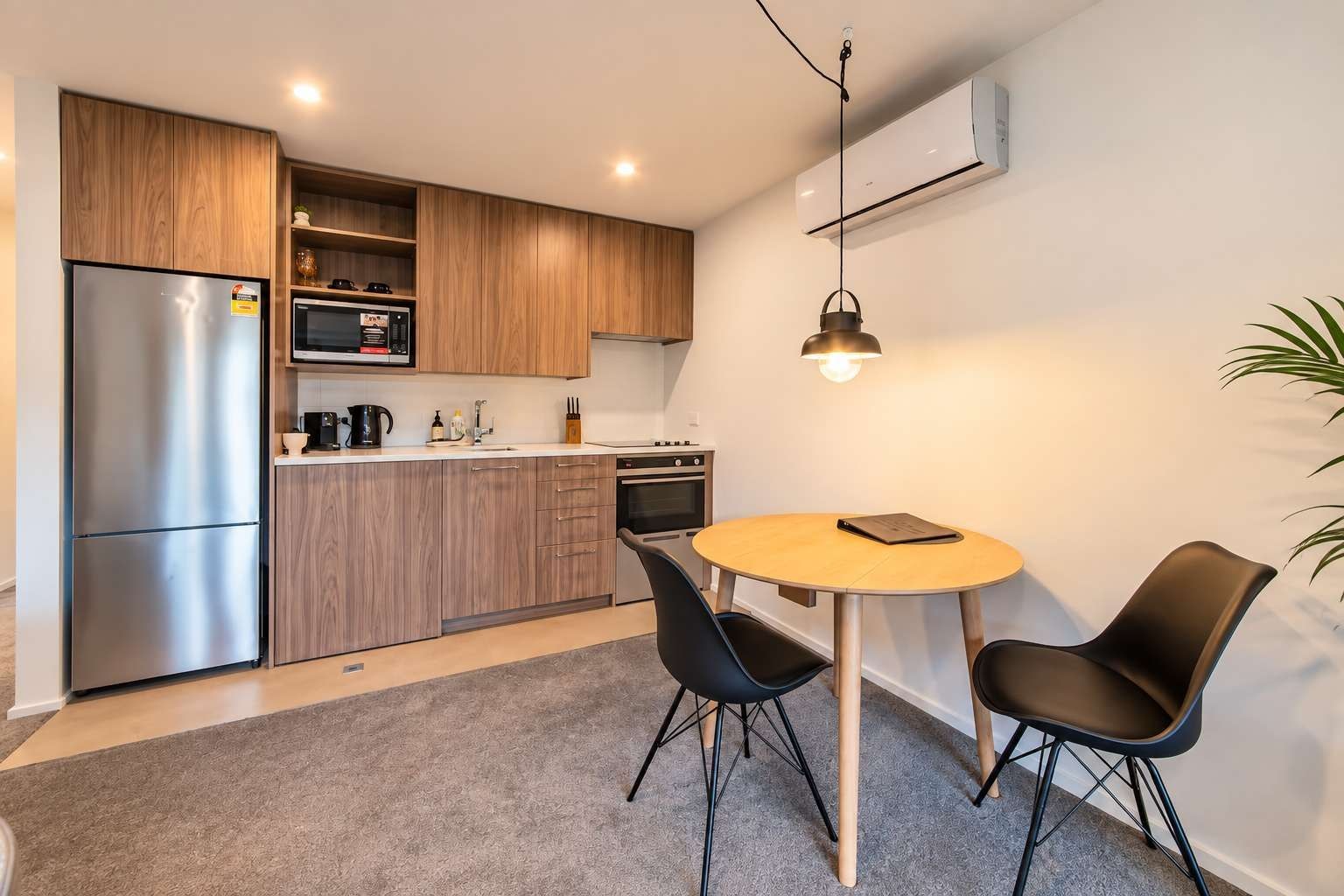 A modern kitchen with a refrigerator, microwave, and stove, a round wooden table with two chairs, and a hanging pendant light.